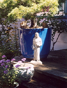 Statue of Saint Fiacre standing by a larg, blue, ceramic pot with a little pine tree in it